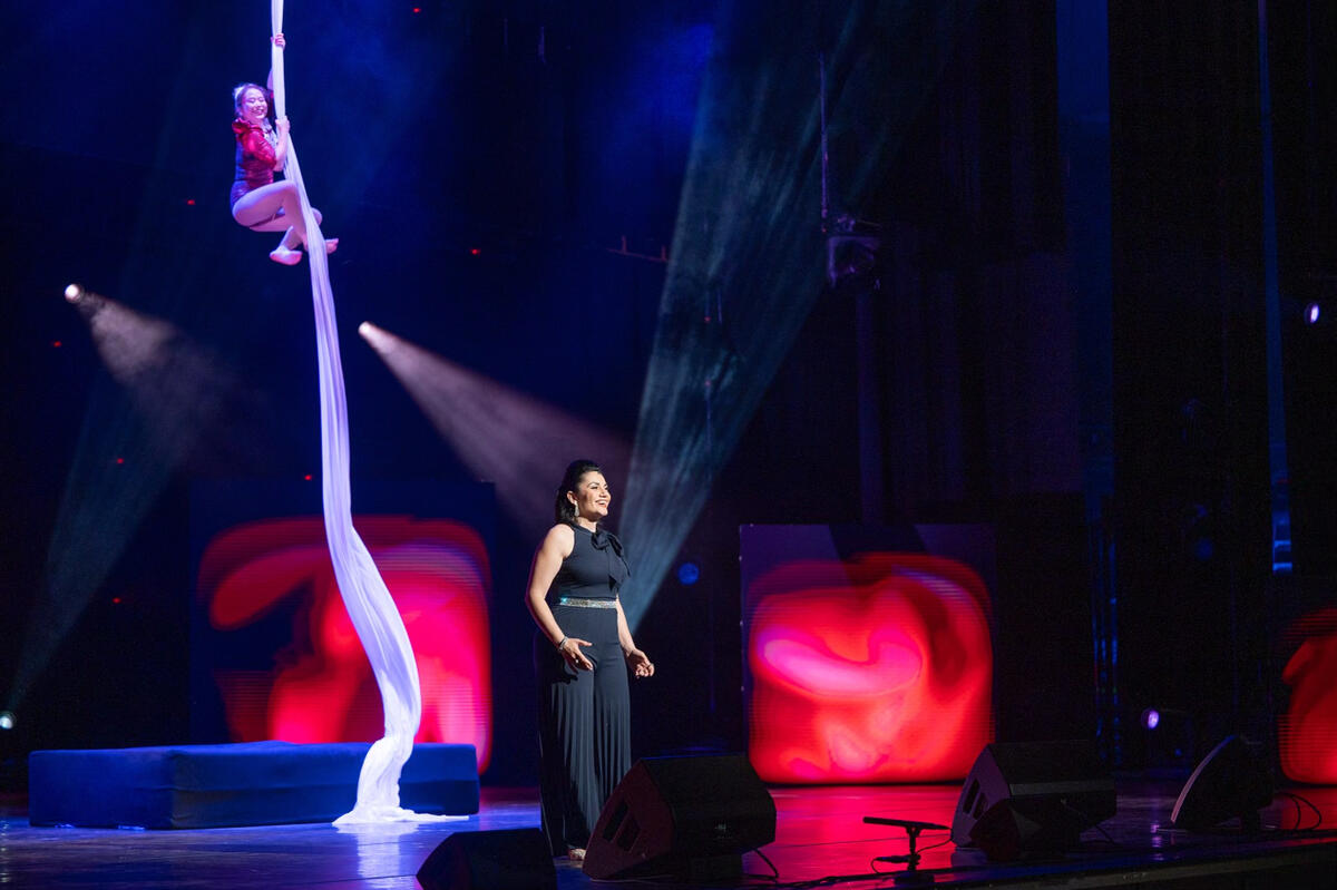 Une chanteuse chante sur scène tandis qu'une acrobate suspendu à un long tissu blanc au plafond exécute une pièce acrobatique en arrière-plan.