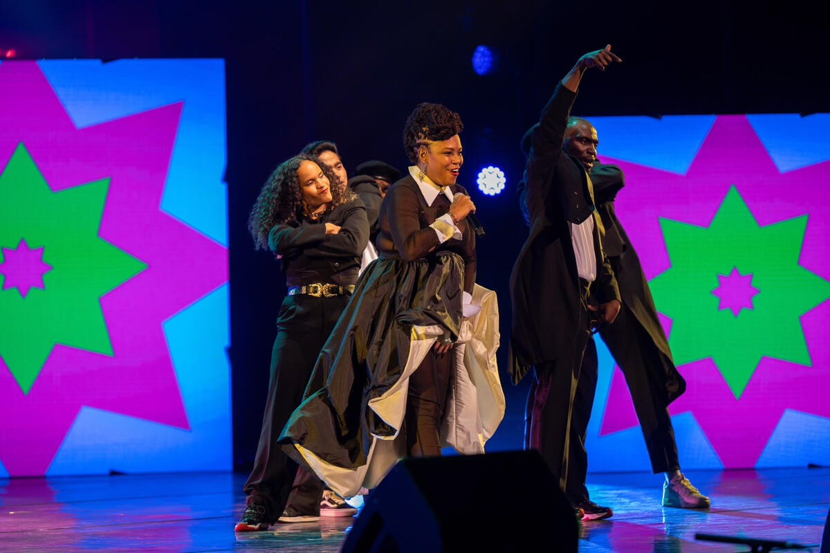 Une chanteuse chante tandis que des danseurs exécutent une routine hip-hop en arrière-plan. La toile de fond est éclairée par un motif très coloré de bleu, vert et violet.
