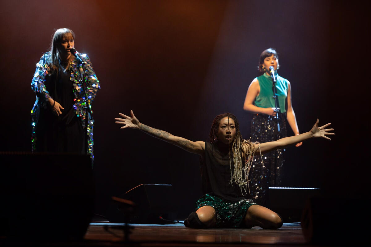 Une artiste exécute un numéro de danse tandis que deux autres chanteurs chantent en arrière-plan.