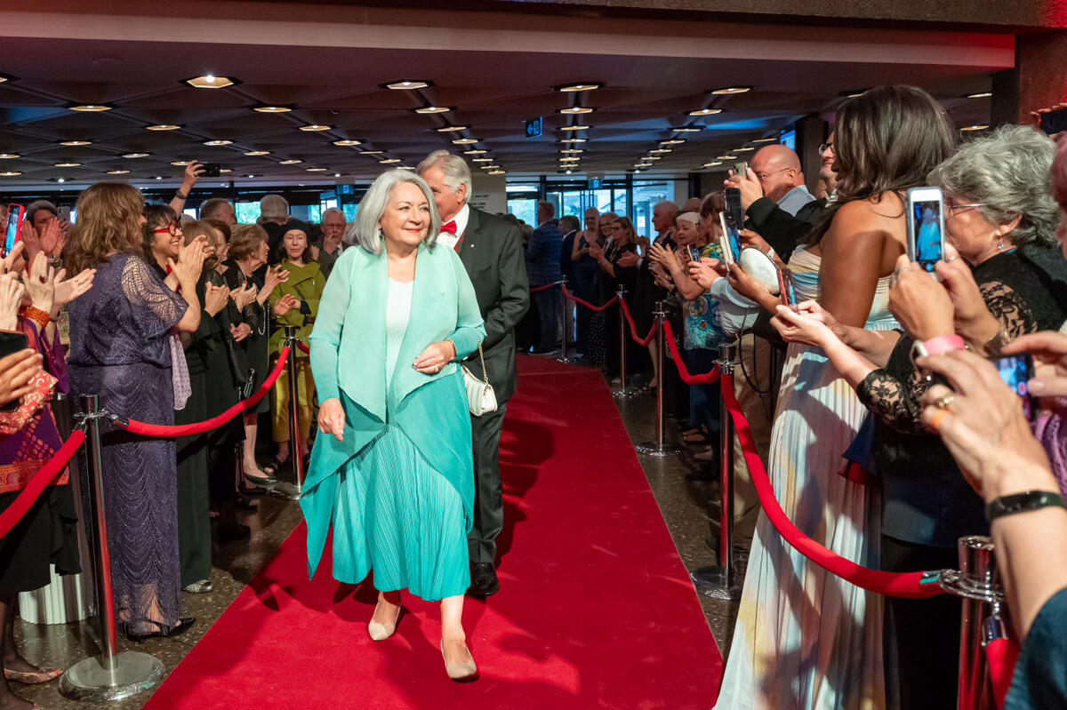 La gouverneure générale Mary Simon marche le tapis rouge, suivie de M. Whit Fraser, lors du gala des Prix du Gouverneur général pour les arts du spectacle. Les personnes tiennent leur téléphone portable pour prendre des photos.