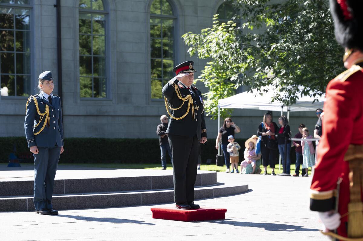 La gouverneure générale Mary Simon salue debout sur une estrade. Un membre de l'Aviation royale canadienne est à sa gauche et des membres de la foule sont visibles à l'arrière-plan. Elle porte également un uniforme de l'Armée canadienne.