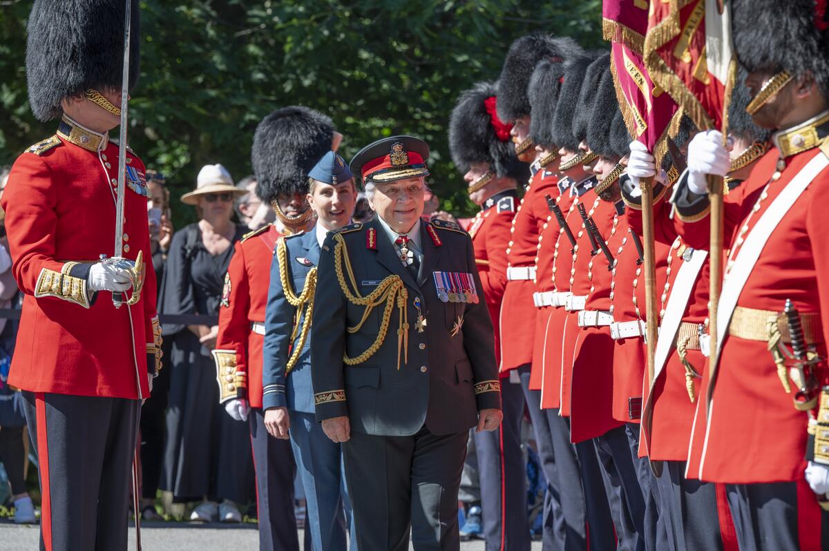 La gouverneure générale Mary Simon inspecte la garde. Un membre de l'Aviation royale canadienne est derrière elle. Elle porte également un uniforme de l'Armée canadienne. Des membres de la garde sont à sa gauche, tous vêtus d'uniformes rouges.