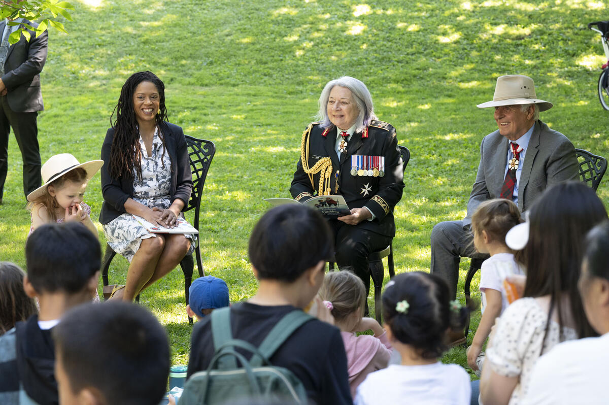 Governor General Mary Simon smiles at the crowd. She is holding a book and is dressed in the Canadian Army uniform. A women sits to her right and Mr. Whit Fraser is seated to her left. There are children in the foreground.