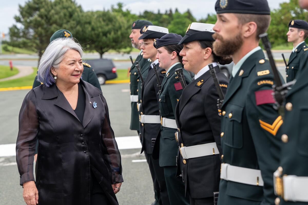 La gouverneure générale Simon passe devant une rangée de personnes en uniforme. Ils sont dehors.