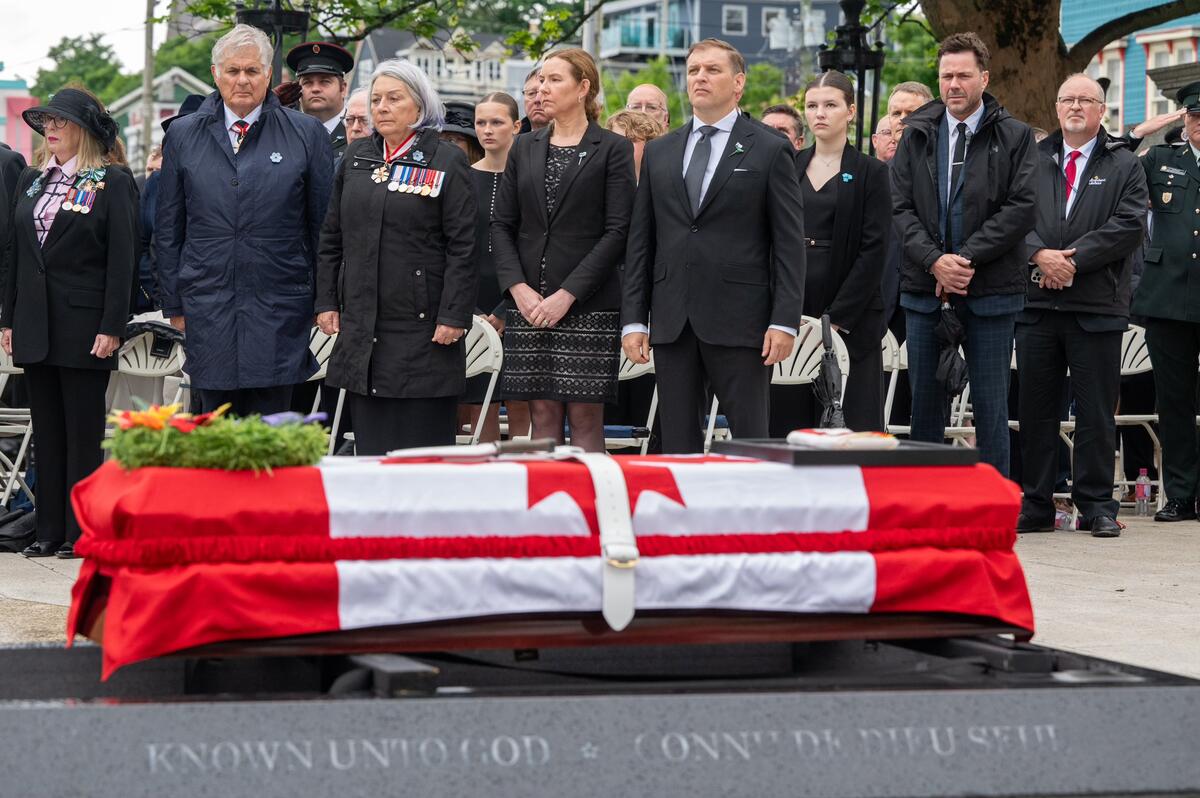 Governor General Simon and Mr. Whit Fraser standing outside with a large group of people at a ceremony. A casket draped with a Canada flag is in the foreground.  