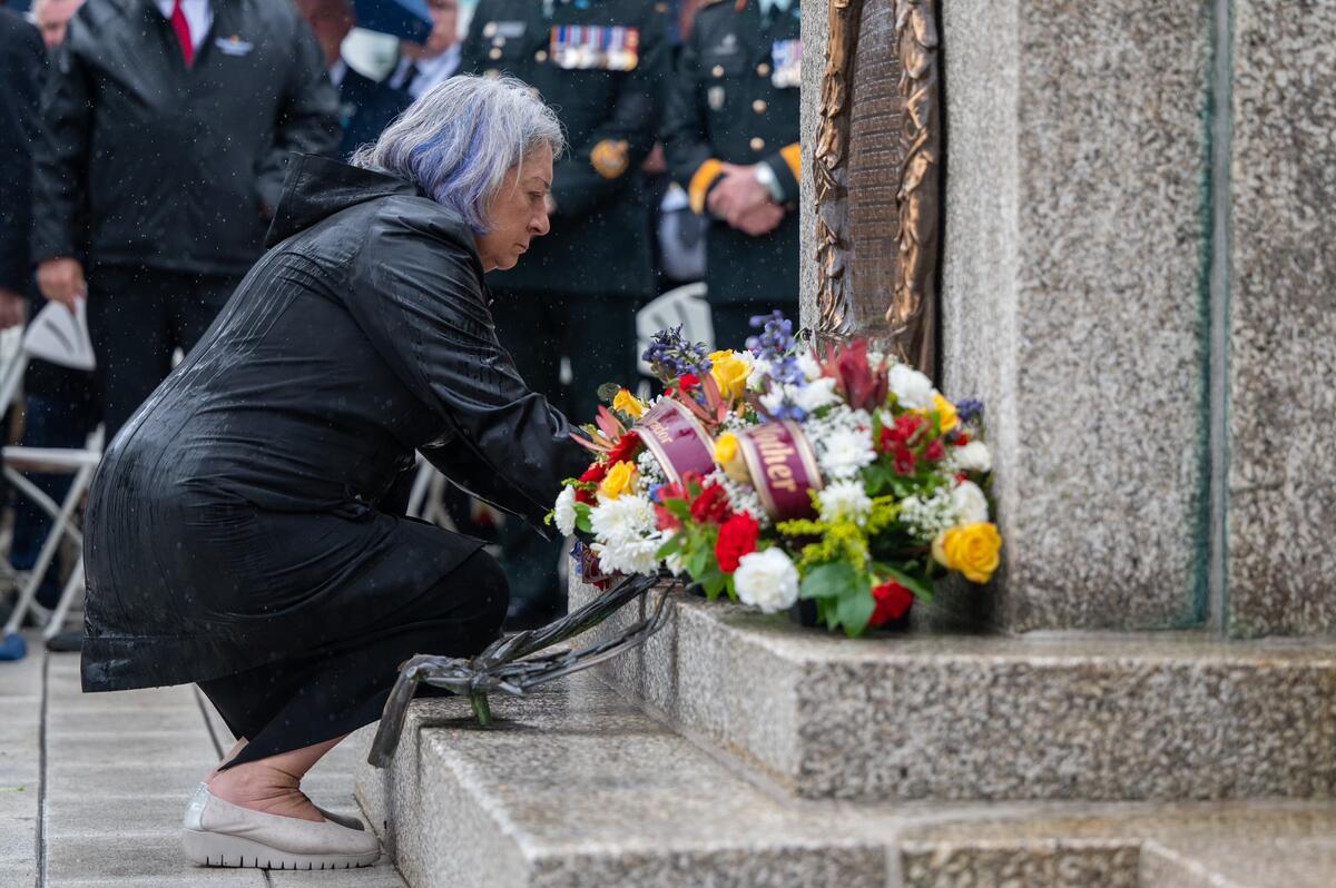 Governor General Simon is crouched down at the foot of a war memorial.  There is a plaque on the wall of the memorial and a bouquet of flowers. 