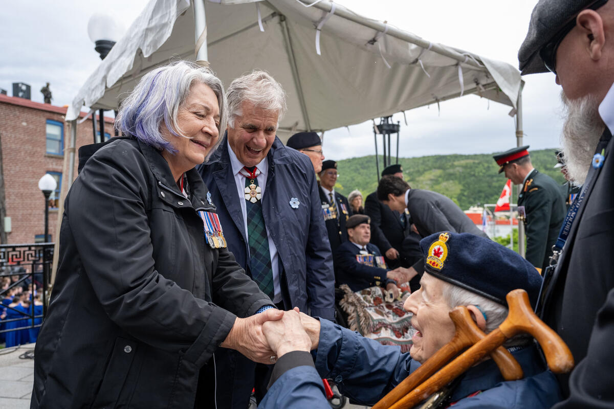La gouverneure générale Simon et M. Whit Fraser s'entretiennent avec un vieil homme en uniforme militaire. La gouverneure générale tient les mains de l'homme. Il est assis et a deux cannes en bois appuyées sur son épaule.