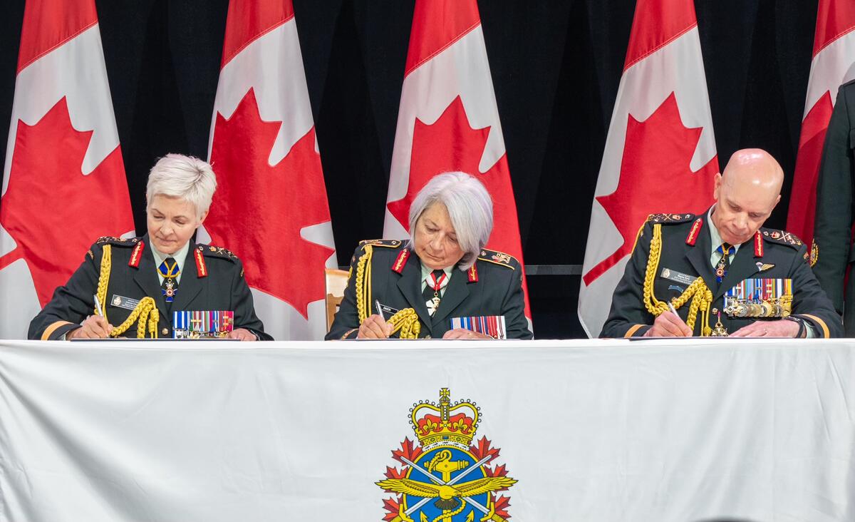 Governor General Mary Simon signs certificates. General Jennie Carignan is sitting to her right and General Wayne Eyre is sitting to her left.