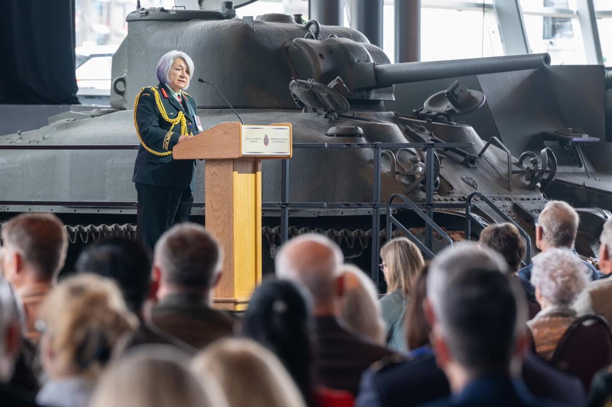 Governor General Mary Simon gives her remarks at a podium. A Sherman tank can be seen in the background and a crowd of people in the foreground.