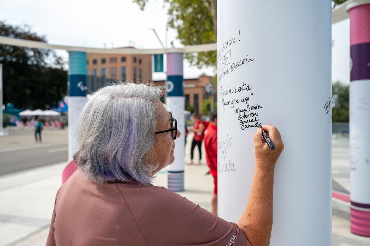 The Governor General of Canada, Mary Simon, writes a message on a white pillar outdoors. The message includes her signature, the phrase "Ajinimata 'Never give up,'" and "Governor General Canada."
