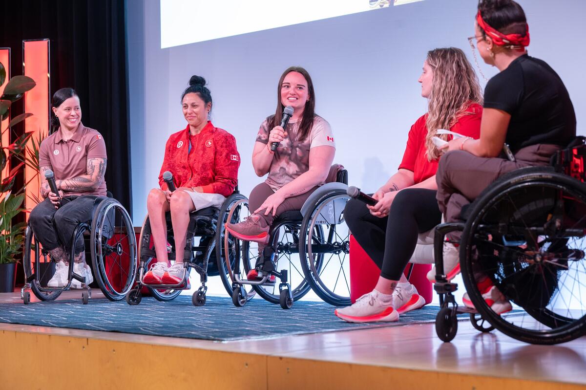 Five people are on a stage, four of whom are in wheelchairs. They are engaged in a conversation; one person is holding a microphone and speaking, while the others listen and smile.