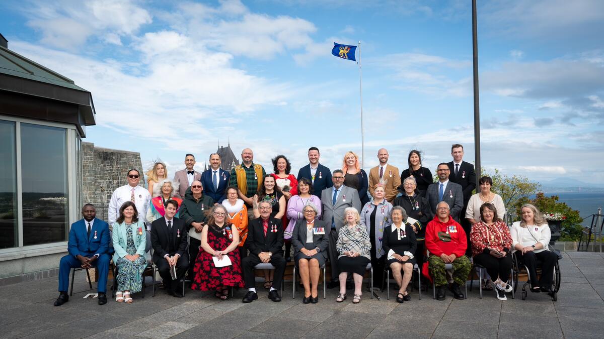 Un groupe de personnes portant des médailles assis sur des chaises à l'extérieur avec la gouverneure générale. Ils sont dehors.