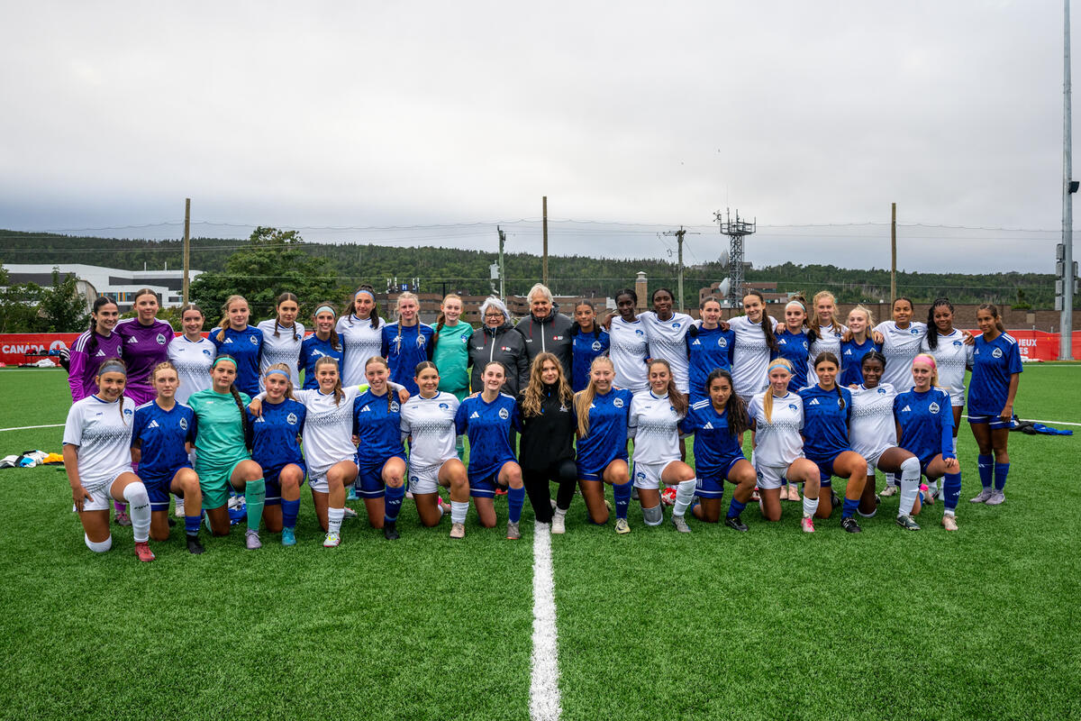 Group photo of a soccer team posing on the field. Governor General Mary Simon and Mr. Whit Fraser stand in the middle of the group.