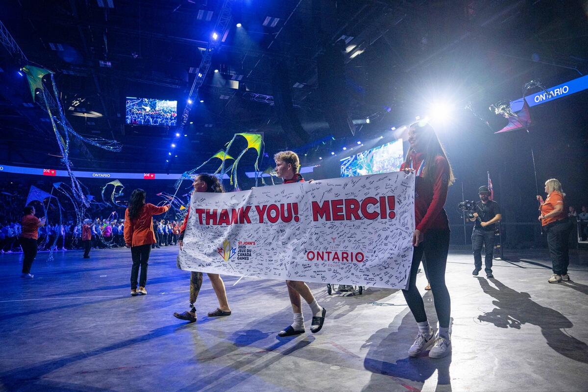 Des athlètes défilant dans un grand stade en brandissant une banderole sur laquelle est inscrit « Merci ! Thank you ! », recouverte de signatures.