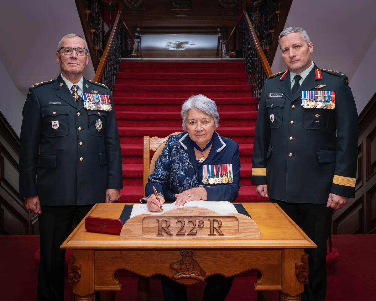 La gouverneure générale Mary Simon assise à un bureau, en train de signer un document, flanquée de deux hommes en uniforme militaire décorés de médailles. Le décor est formel, avec un escalier recouvert d'un tapis rouge.