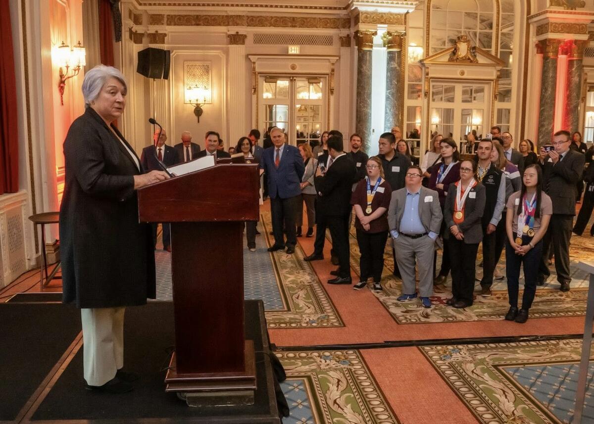 Governor General Mary Simon speaking at a podium in an ornate room filled with attentive people. The setting is formal, with decorative carpets and elegant lighting.