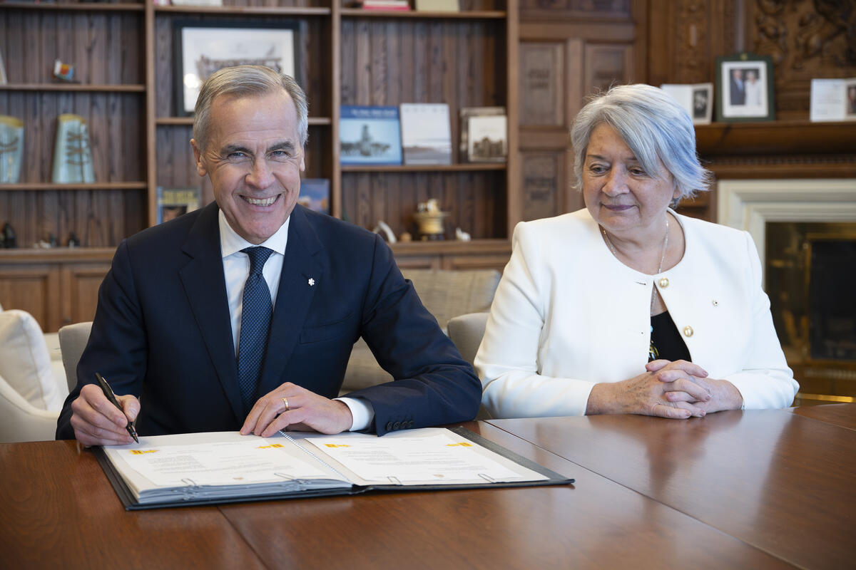 Prime Minister Mark Carney and Governor General Mary Simon are sitting at a large wooden table. Mark Carney is signing a document.