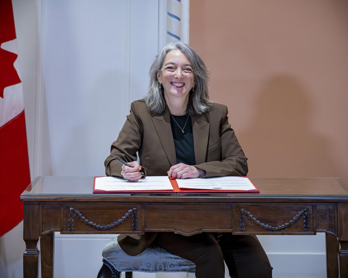 Julie Aviva Dabrusin sitting at a wooden desk signing a document. There is a Canada flag to his right.