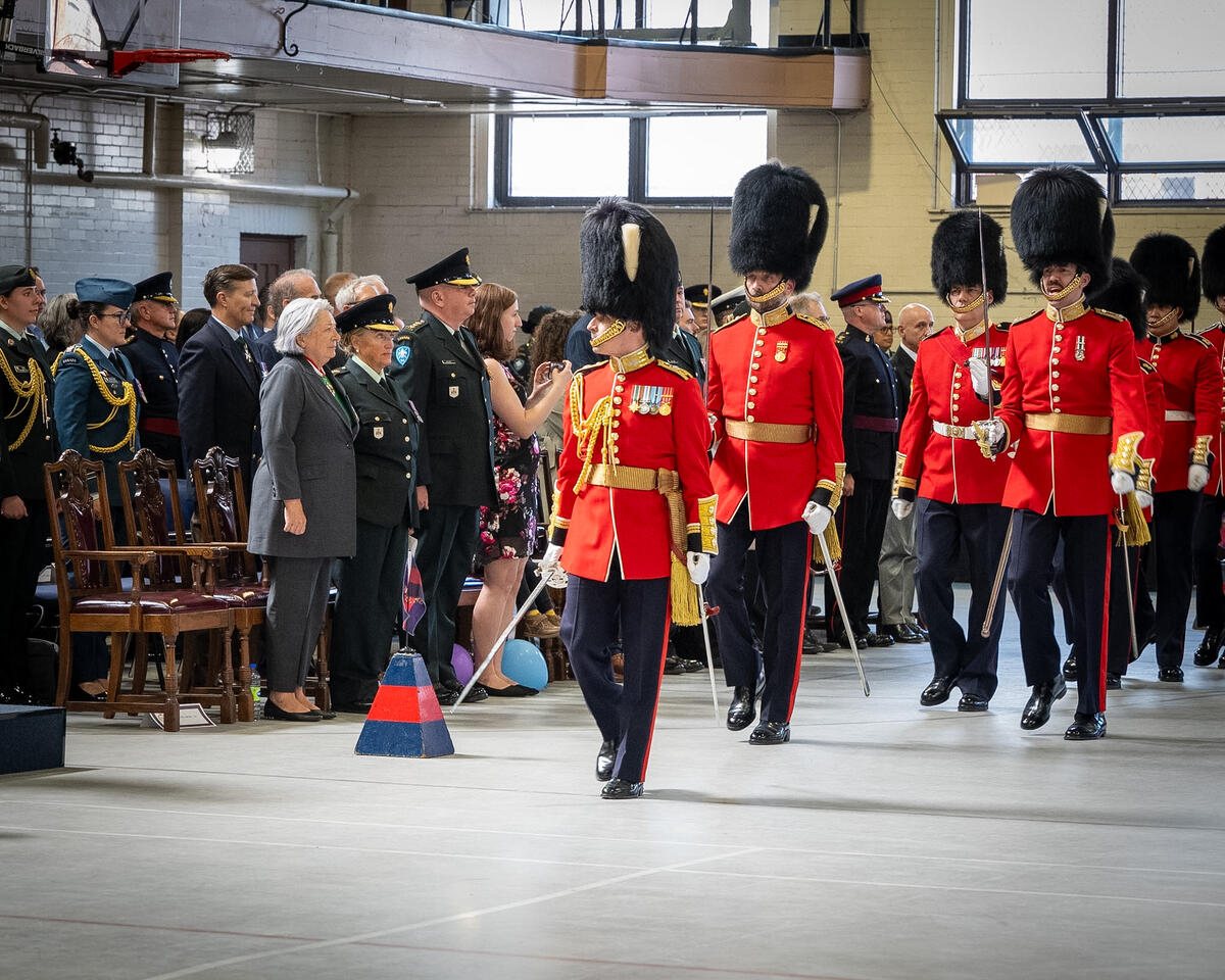 La gouverneure générale Mary Simon se tient debout avec un groupe de personnes tandis que les Canadian Grenadier Guards défilent. Ils se trouvent dans un grand gymnase.