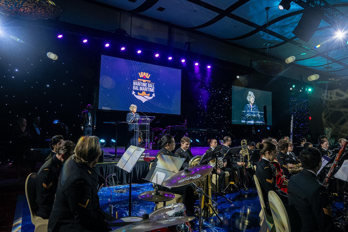 La gouverneure générale Arbour se tient au podium lors du Bal des Maritimes, tandis qu’un orchestre, vêtu d’uniformes de la marine et jouant de ses instruments, l’écoute.