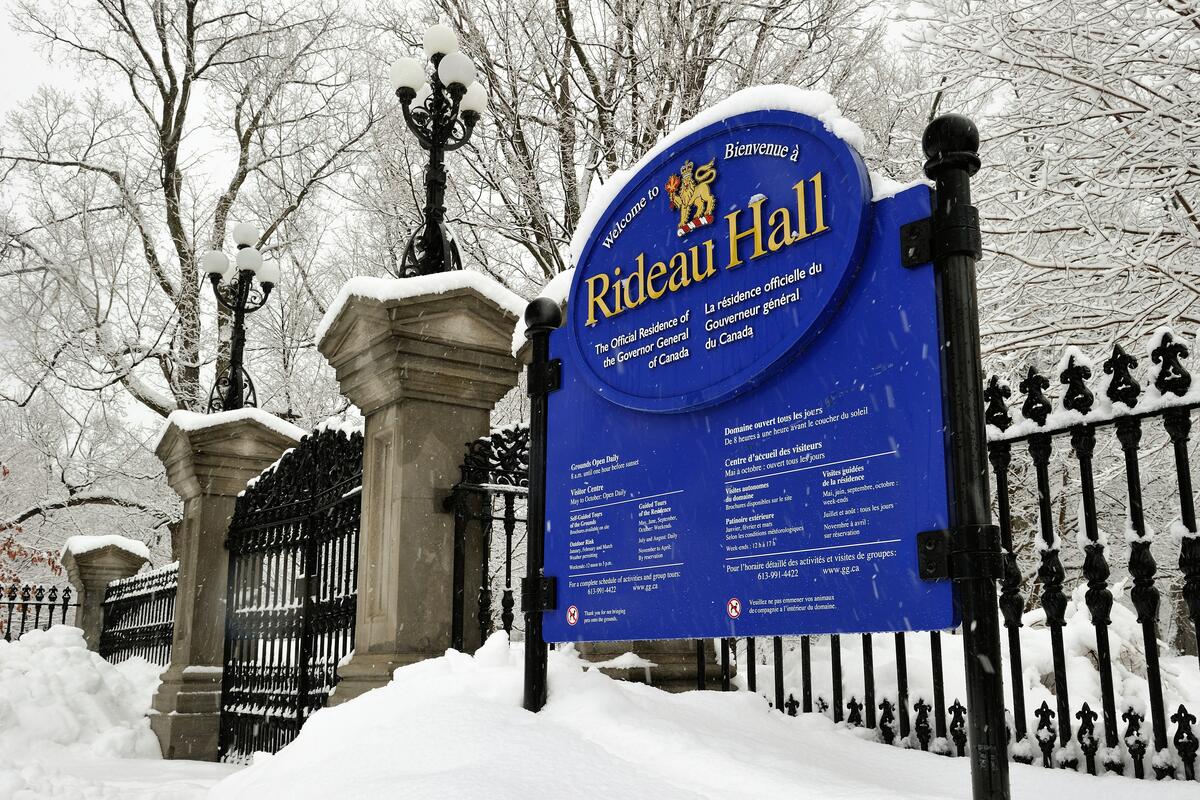 Panneau recouvert de neige indiquant Rideau Hall, la résidence officielle du gouverneur général du Canada. Avec en arrière-plan une clôture en fer forgé et des arbres en hiver.