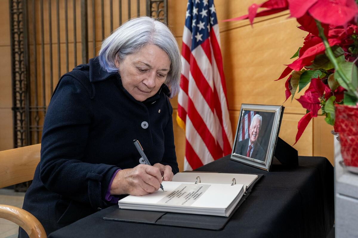Une femme aux cheveux blancs est assise à une table recouverte d'un tissu noir et signe un livre à côté d'une photo encadrée de l'ancien président américain Jimmy Carter.