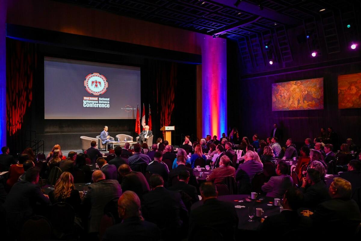 Une grande salle sombre, avec une scène éclairée sur laquelle deux hommes sont assis sur des fauteuils. Sur un écran derrière eux est écrit .First annual National Indigenous Defence Conference » sous un logo rouge, orange et blanc avec un oiseau rouge.