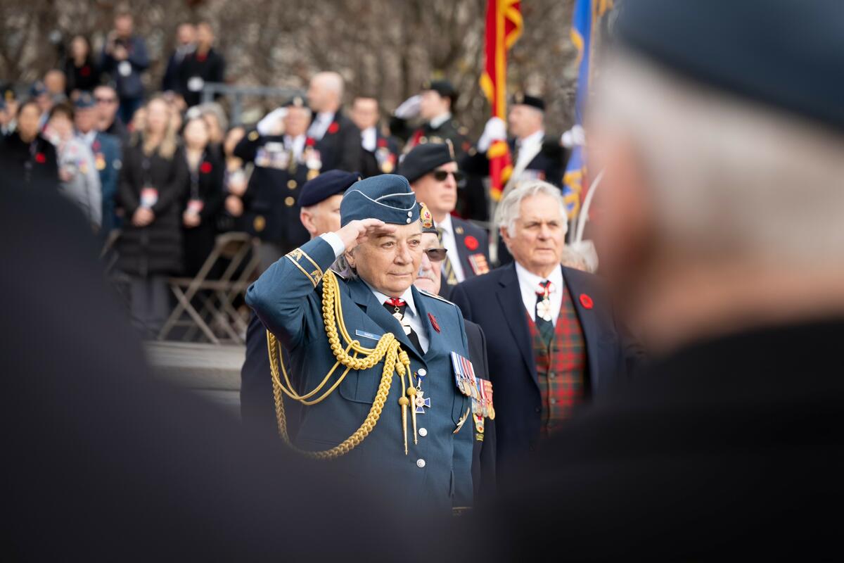 Governor General Mary Simon, wearing the Air Force uniform, is saluting. There is a crowd of people behind her. Her husband Whit Fraser is on her right.