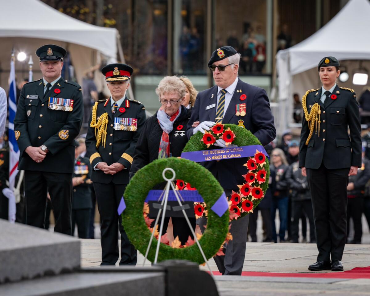 A military member with grey hair is holding a wreath of flowers with a ribbon that says "The Mothers of Canada". A lady with glasses, wearing a light blue and red scarf around her neck, bowing her head, is next to him. they are surronded by miltrary membe