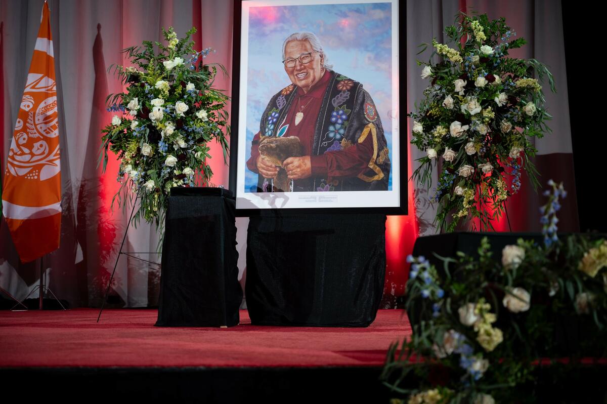 Portrait peint de Murray Sinclair sur une scène recouverte d'un tapis rouge, entouré de fleurs funéraires.