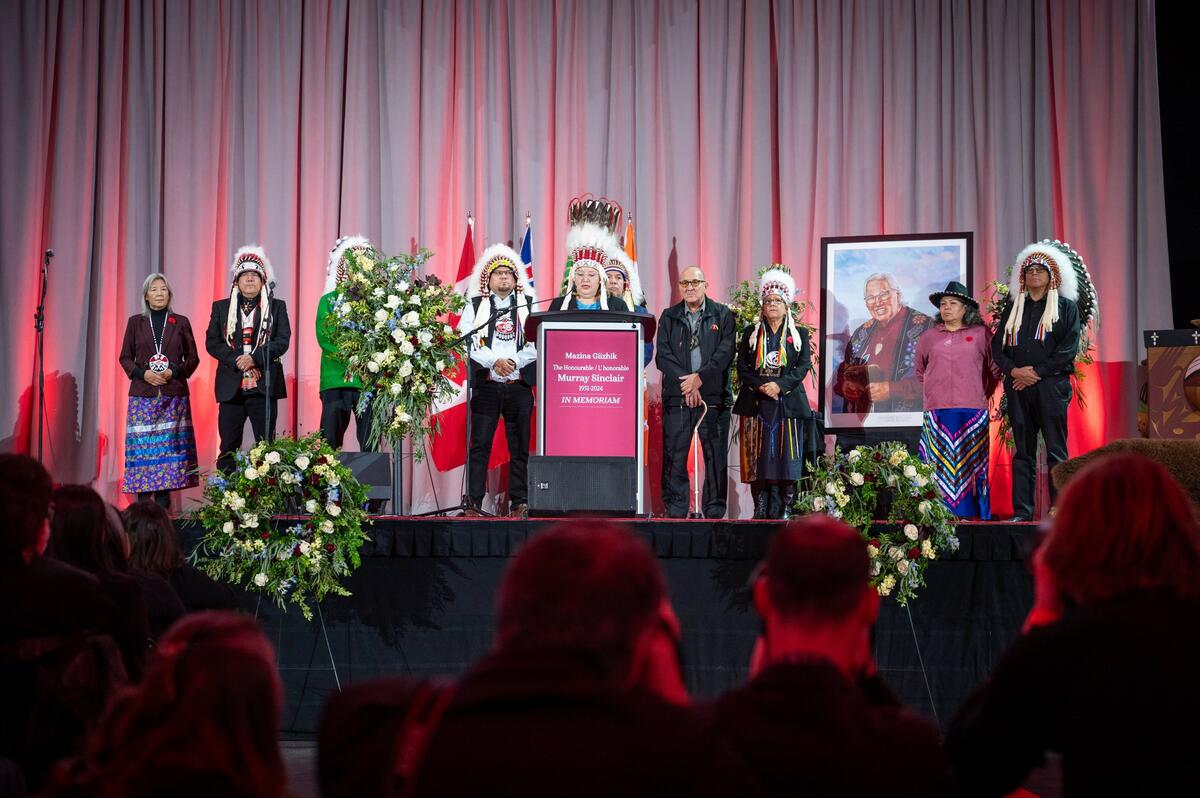Un groupe d'autochtones, dont certains portent des vêtements traditionnels, se trouve sur une scène. L'un d'eux parle sur un podium. On voit derrière eux le portrait peint de Murray Sinclair.