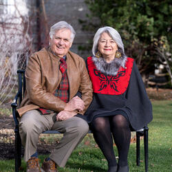 Governor General Mary Simon and Mr. Whit Fraser are sitting on a park bench, outside, smiling and holding hands, on a sunny fall day.