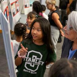A young girl in a green "Stanley Public Schools" t-shirt explains a project on a display board to an older woman in glasses, in a lively classroom setting.