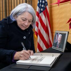 A white haired lady is sitting at a table covered with a black cloth, signing a book next to a framed photo of former U.S. President Jimmy Carter.