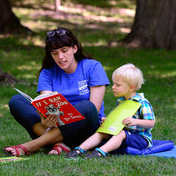 Une femme lit un livre à un petit garçon. Ils sont assis sur une couverture bleue sur l'herbe à l'extérieur.