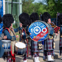 Drummers and pipers wearing military uniforms are performing outside. A large drum reads, “100 RCAF.”