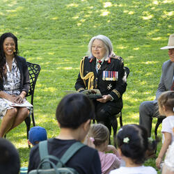 Governor General Mary Simon smiles at the crowd. She is holding a book and is dressed in the Canadian Army uniform. A women sits to her right and Mr. Whit Fraser is seated to her left. There are children in the foreground.