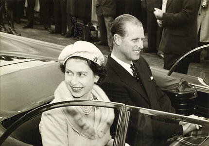 La reine et le duc d’Édimbourg sourient à la foule alors qu’ils sont à bord d’un véhicule décapotable. La photo est en noir et blanc.