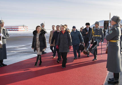 La gouverneure générale marche sur un tapis rouge qui mène à l'escalier d'un avion à l'arrière-plan.  Elle est entourée de représentants officiels.  Des gardes armés tapissent le tapis rouge.