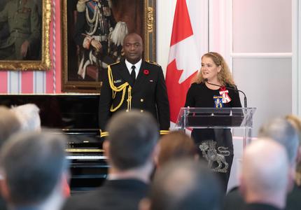 La gouverneure générale prononce une allocution debout devant un podium.  Derrière elle se trouve son aide de camp en uniforme de la Marine ainsi que le drapeau canadien.  