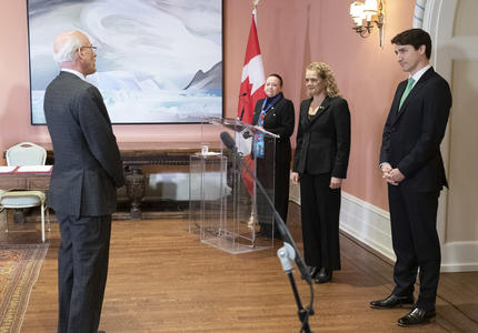 Michael Wernick, greffier du Conseil privé, se tient devant la gouverneure générale Julie Payette et le premier ministre Justin Trudeau.   Dans le coin de la salle, le maître de cérémonie se tient derrière un podium.