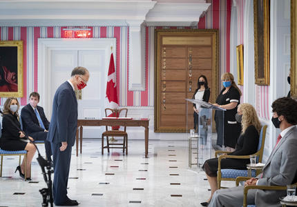 Un homme en costume est debout devant la gouverneure générale qui est assis. Il la reconnaît d'un signe de tête. Le Premier ministre regarde sur le côté.