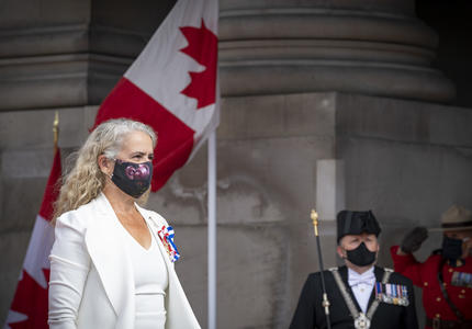 Une femme vêtue d'un costume blanc avec un masque noir est en train de marcher. Un homme en uniforme noir et un agent de la Gendarmerie royale du Canda portant un uniforme rouge saluent en arrière-plan. Il y a également deux drapeaux canadiens.