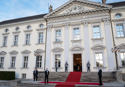 Grand bâtiment blanc de style victorien avec un tapis rouge à l'extérieur. La porte d'entrée est ouverte. Il y a trois hommes en uniforme militaire et trois hommes en costume.