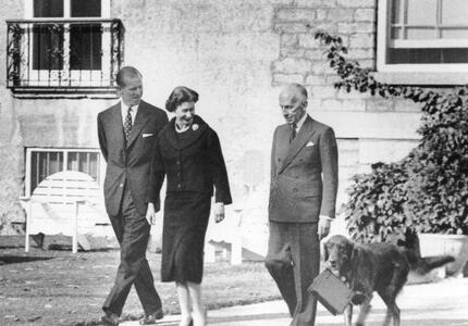 Photo en noir et blanc de Sa Majesté la Reine et du duc d’Édimbourg marchant à l’extérieur de Rideau Hall avec le gouverneur général Massey. Un chien marche à leurs côtés en transportant dans sa gueule le sac à main de la Reine. 