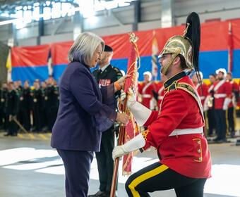 La gouverneure générale Mary Simon porte un costume violet et sourit à un membre des Forces armées canadiennes alors qu'elle lui remet le guidon. Le membre des Forces armées canadiennes porte un uniforme rouge, des gants blancs et un casque. 