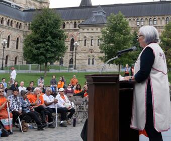 Governor General Mary Simon is standing at a podium. People are seated in front of her. Many people are wearing orange shirts. There is a large stone building in a background.