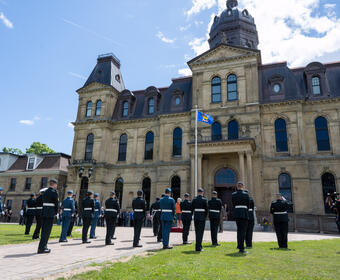  A group of people in military uniforms standing in rows and facing large stone building. The viceregal flag is flying on a pole.