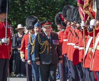 Governor General Mary Simon inspects the guard. A member of the Royal Canadian Air Force is behind her. She is also wearing a Canadian Army uniform. Members of the guard are to her left, all wearing red uniforms.