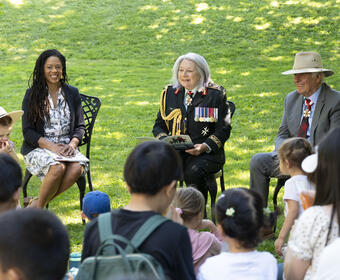 Governor General Mary Simon smiles at the crowd. She is holding a book and is dressed in the Canadian Army uniform. A women sits to her right and Mr. Whit Fraser is seated to her left. There are children in the foreground.